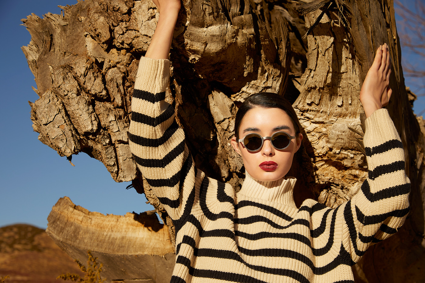 Model in striped sweater and sunglasses posing by tree trunk in desert editorial by Vikram Pathak