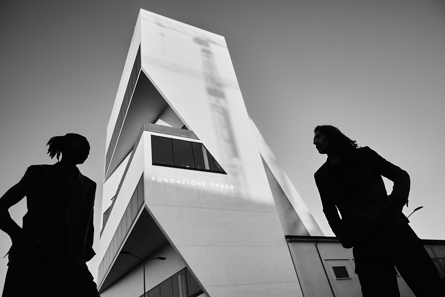 Two male models photographed in silhouette against the architecture of Fondazione Prada Milan by Vikram Pathak.