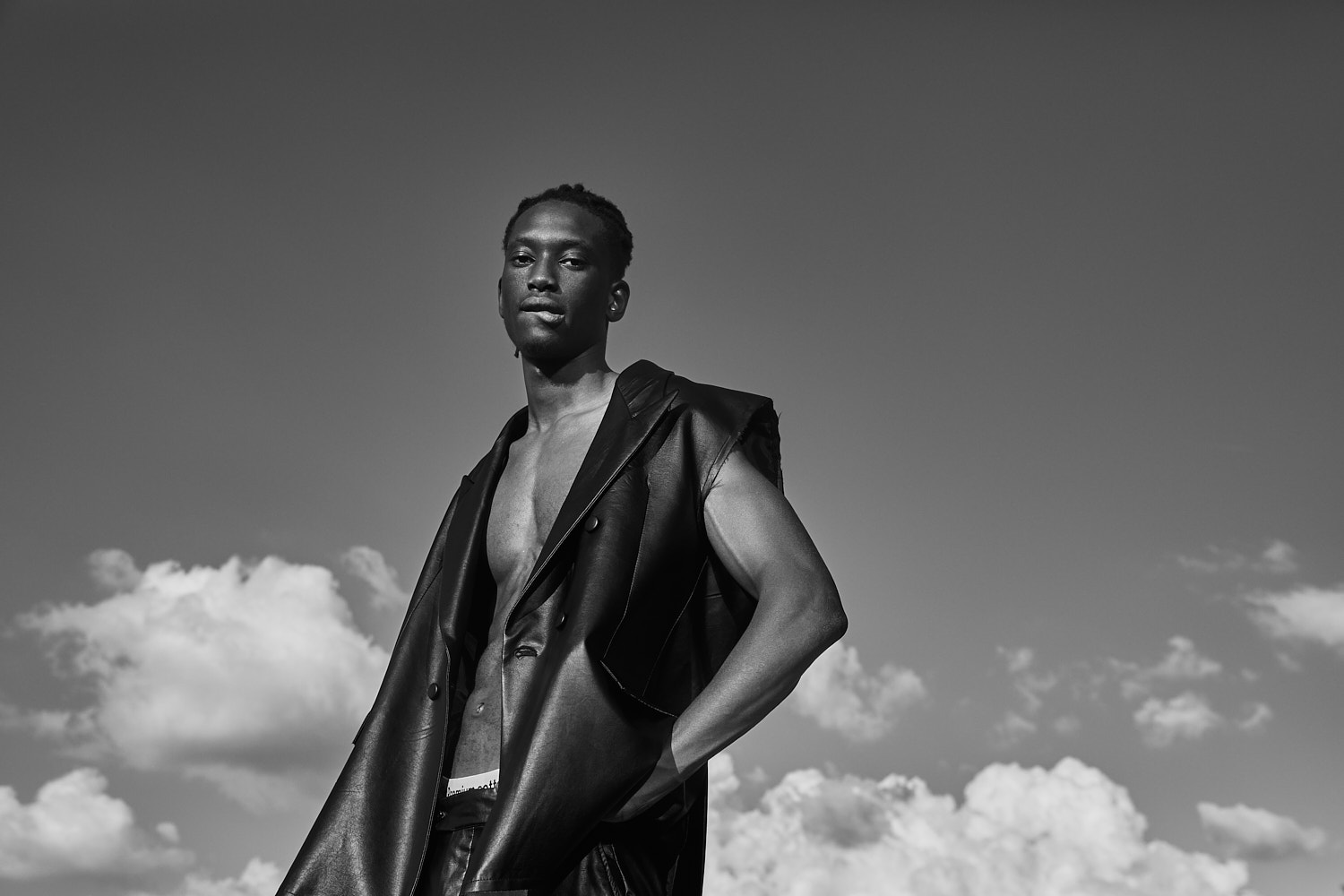 Male model posing in sleeveless black suit under blue sky and clouds, photographed by Vikram Pathak.