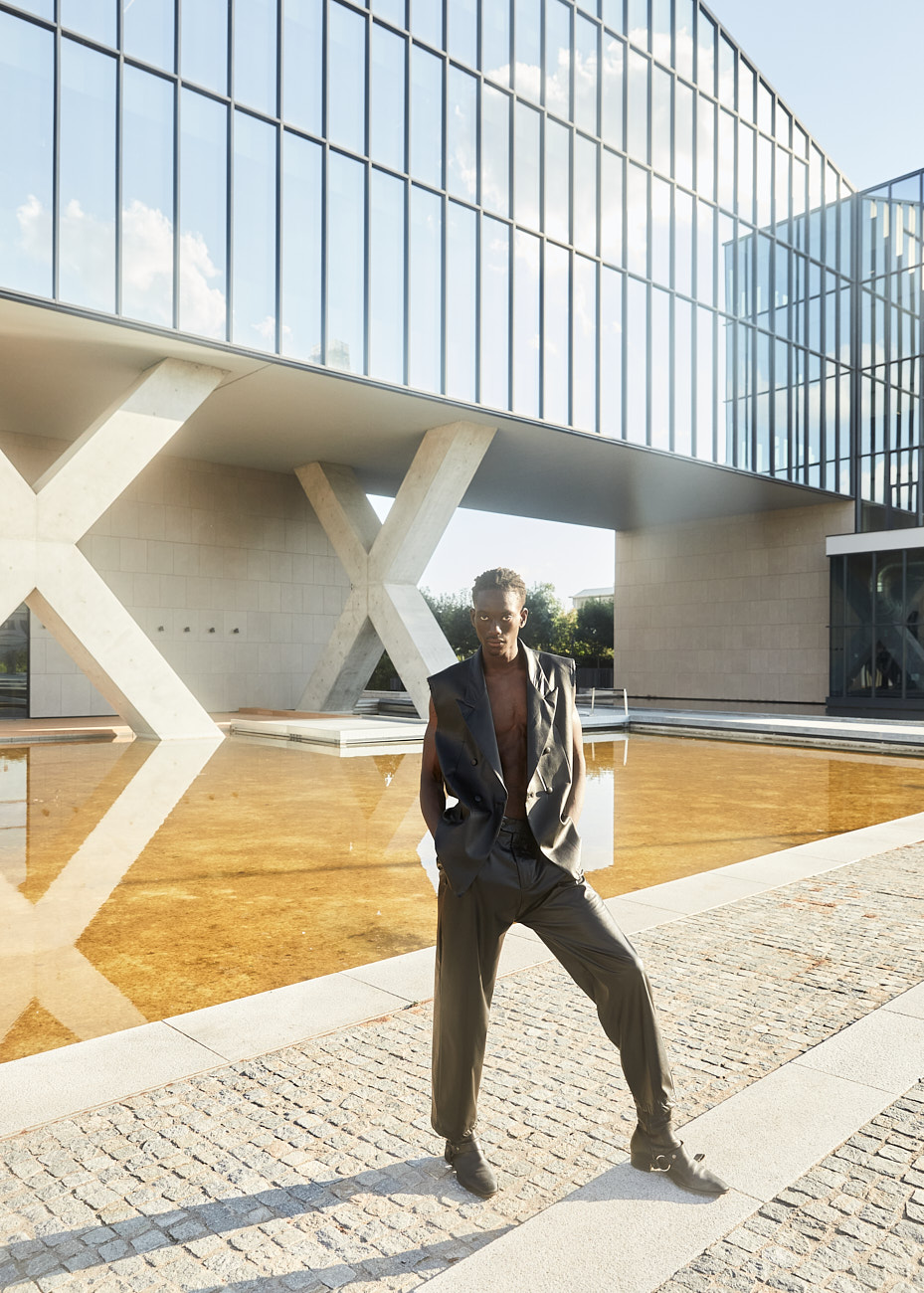 Full-length portrait of male model in sleeveless black suit standing by reflective pool at Prada Foundation Milan.