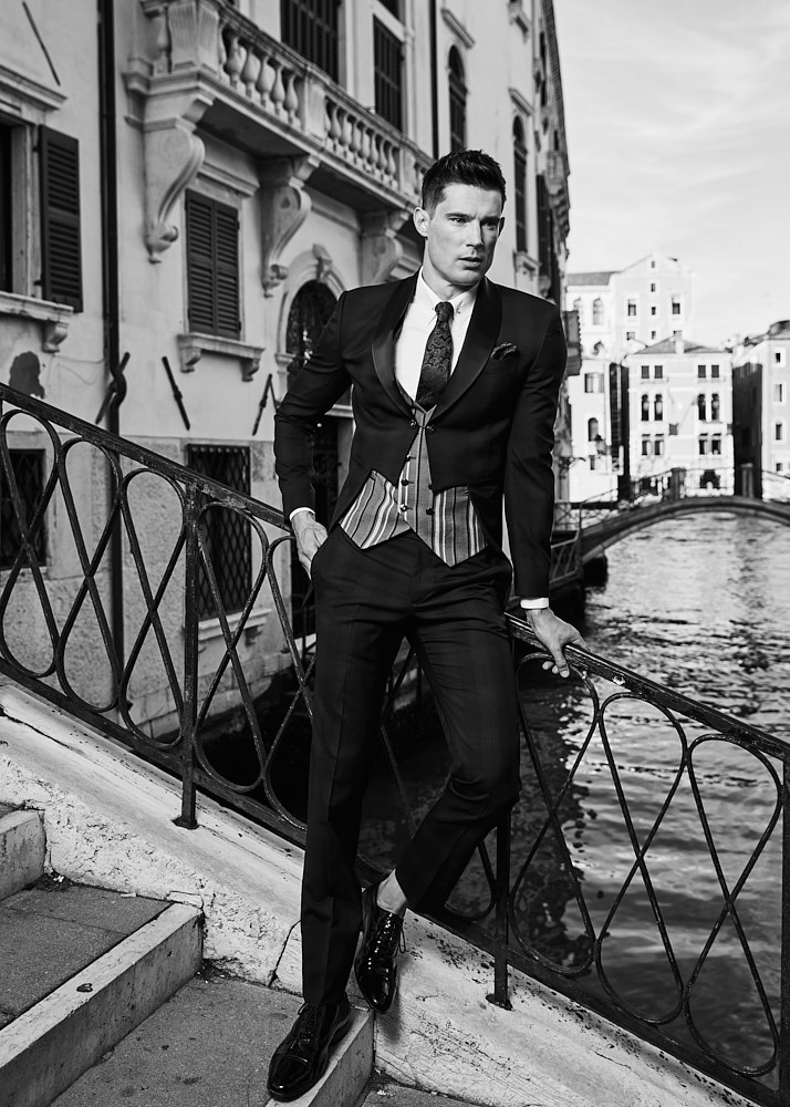 Male model in black tuxedo posing on Venetian bridge overlooking canal, editorial fashion photo