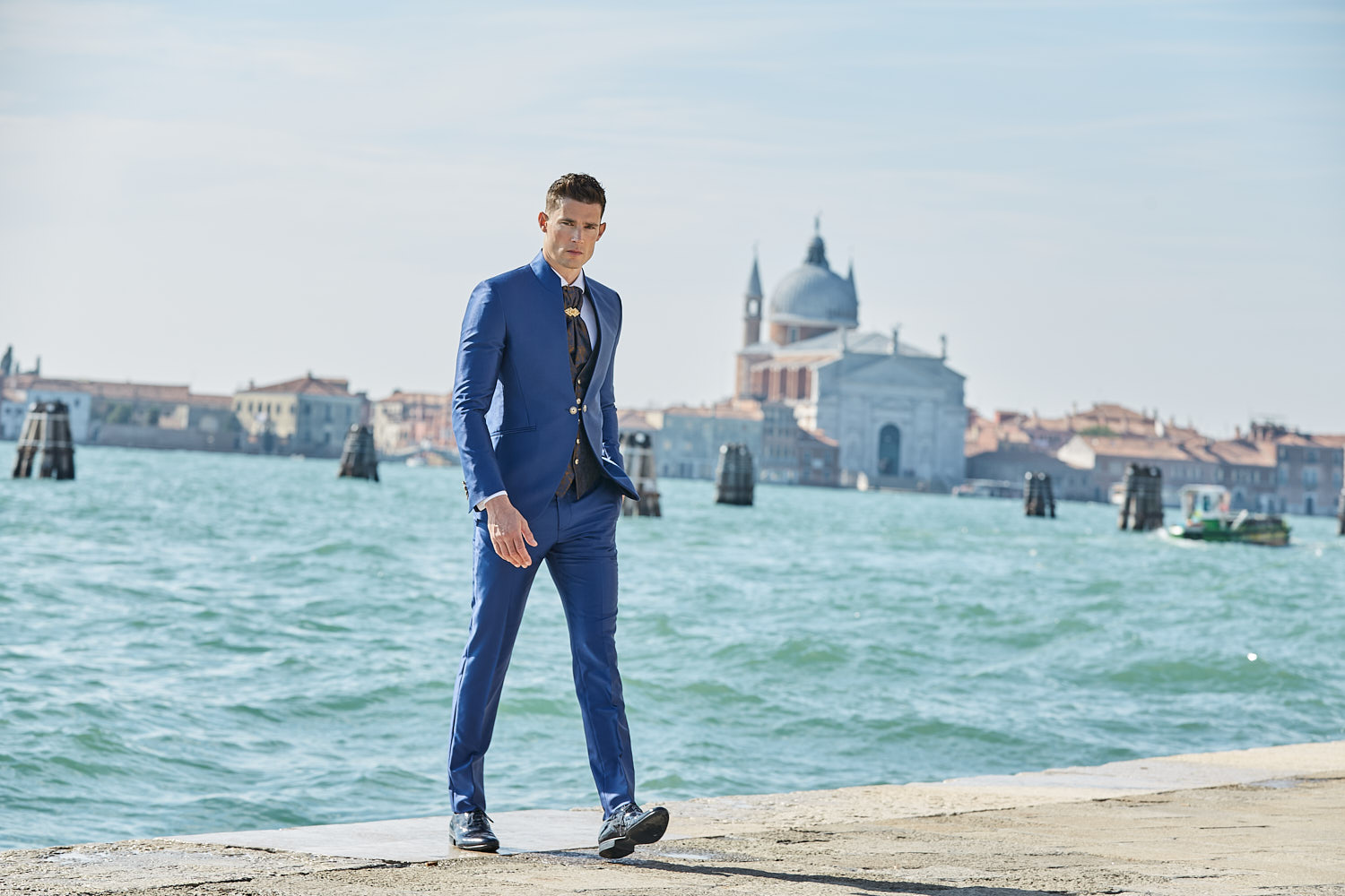 Male model in tailored blue suit walking by Venice waterfront with domes in background