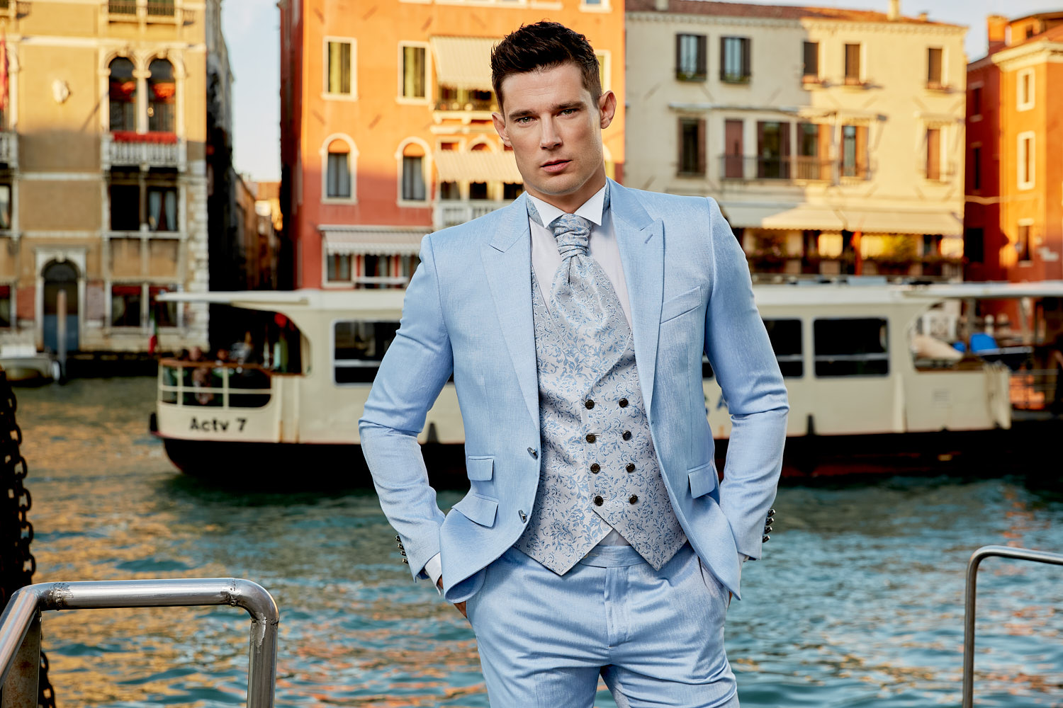 Male model in light blue three-piece suit posing near Venice water taxi on Grand Canal