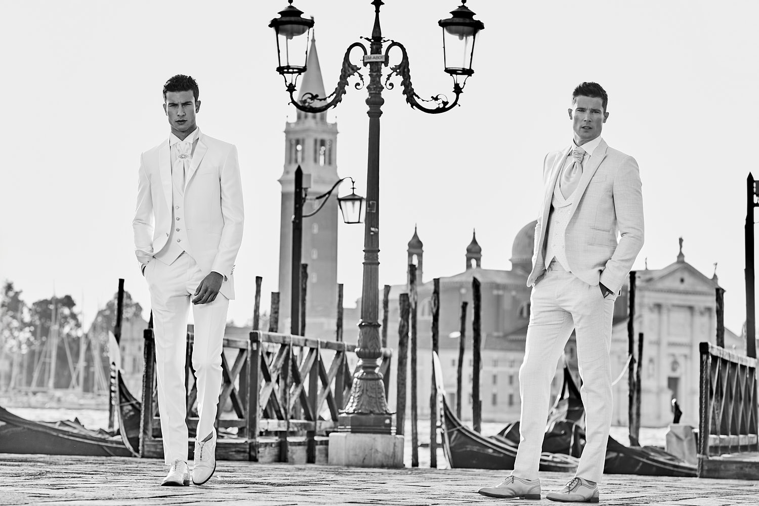 Two male models in white suits posing by gondolas with historic architecture in Venice