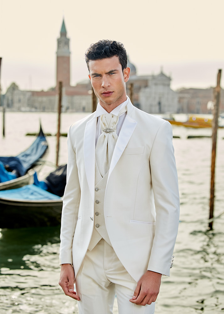 Male model in cream suit and silk tie standing near gondolas in Venice fashion photoshoot