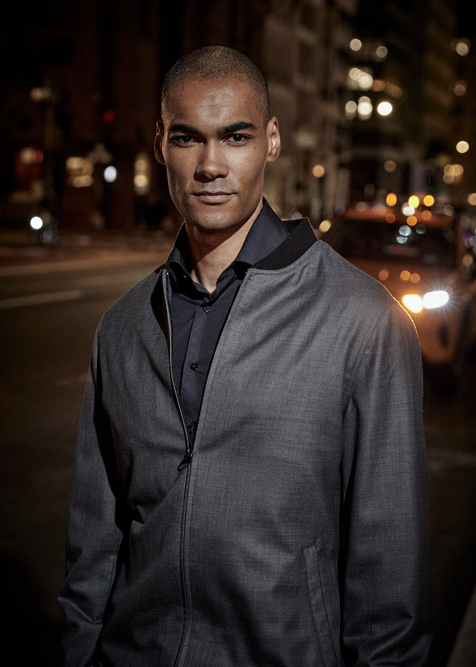Male model wearing a grey zip-up jacket with black shirt at night in NYC with taxi cabs in the background