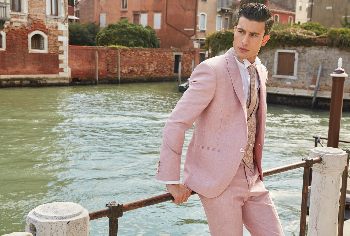 Editorial portrait of a man in a light pink suit posing by a Venetian canal.