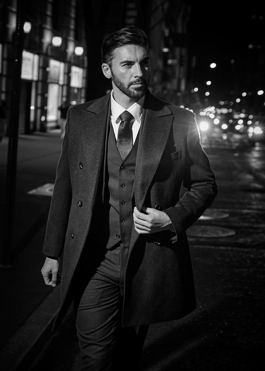 Man in long tailored overcoat walking at night in NYC during black and white editorial fashion shoot by Vikram Pathak